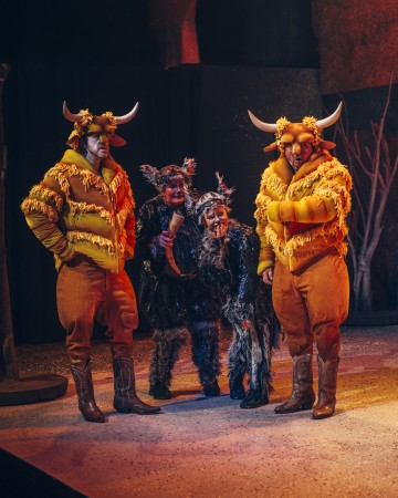 Lavalla seisoo neljä teatteripukuista henkilöä. Kahdella on keltaiset sarvipäiset härkäasut ja puskurihousutakit, ja kahdella muulla on karvaiset mustat ja harmaat eläinpuvut. Taustalla on tummia oksia ja kiviä.