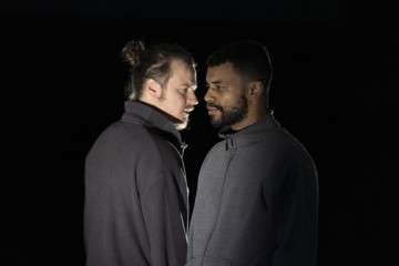 Kaksi harmaatakkista miestä seisoo lähekkäin pimeässä ympäristössä. Toinen mies katsoo rauhallinen ilme kasvoillaan eteenpäin, kun taas toinen, hieman takana seisova mies katsoo häntä vakavasti tai intensiivisesti.