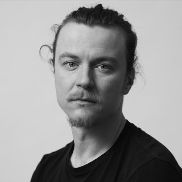 Henkilö, jolla on vaalea iho, vaaleat silmät ja taakse sidotut pitkät hiukset, päällään musta paita, katsoo vakavana kameraan vaaleaa taustaa vasten.