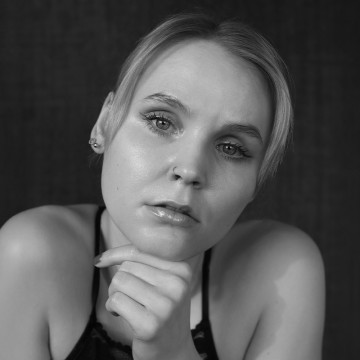 Nuori nainen, jolla on lyhyet vaaleat hiukset, katsoo mietteliäästi kameraan ja nojaa leuka käteensä. Hänellä on yllään spagettinauhatoppi ja pienet korvakorut. Kuva on mustavalkoinen ja siinä on tumma tausta.