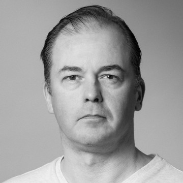 Keski-ikäinen mies, jolla on lyhyet tummat hiukset ja vaalea paita, katsoo suoraan kameraan neutraalilla ilmeellä vaaleanharmaata taustaa vasten.