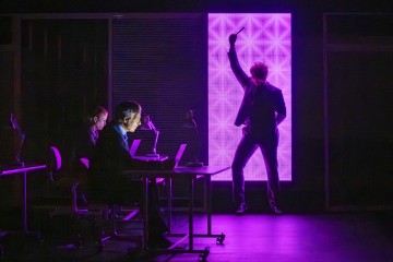 Kolme ihmistä on pimeässä huoneessa; kaksi istuu työpöydän ääressä kannettavien tietokoneiden kanssa, kun taas yksi seisoo hehkuvan vaaleanpunaisen geometrisen valopaneelin edessä ja kohottaa kättään dramaattisesti.