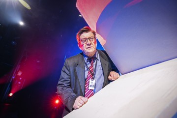 Pukuun ja raidalliseen solmioon pukeutunut mies seisoo dramaattisessa valaistuksessa ja katsoo alaspäin valkoisen kulmikkaan rakenteen luona, jonka taustalla on punaisia ja sinisiä valoja. Hänellä on silmälasit ja henkilökortti kaulassaan.