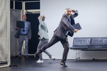 Pukumies juoksee innoissaan kansio kädessään hymyillen ja vilkuttaen, kun kaksi muuta ihmistä kannettavien tietokoneiden kanssa seisoo taustalla sinisen sohvan vieressä modernissa toimistoympäristössä.