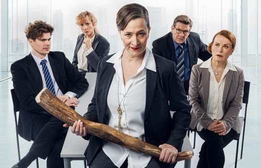 Viisi liikemiestä istuu toimistossa. Edessä seisoo itsevarma nainen, jolla on kädessään suuri puukeppi, kun taas muut hänen takanaan näyttävät yllättyneiltä tai huolestuneilta. Ikkunoista näkyy kaupungin rakennuksia.