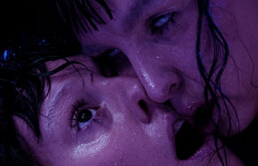 Kaksi ihmistä lähekkäin, heidän kasvonsa painautuvat yhteen dramaattisessa violetissa valaistuksessa. Toisen henkilön silmät ovat auki, kun taas toinen näyttää kuiskaavan tai hengittävän suunsa lähellä. Molemmat vaikuttavat intensiivisiltä ja tunteikkailta.