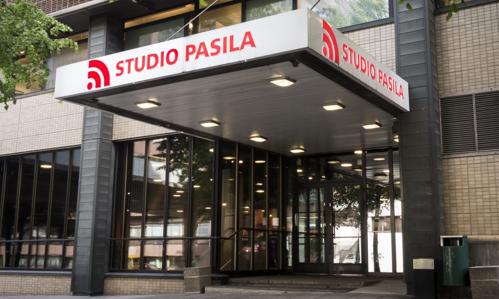 Studio Pasilan sisäänkäynti, suuri valko-punainen kyltti lasiovien yläpuolella. Rakennuksen ulkopinnassa on vaaleat laatat, tummat ikkunanpuitteet ja lähellä pieni vihreä roskakori.
