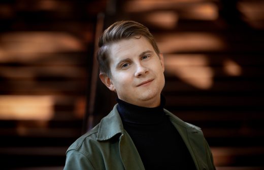 Lyhyet vaaleanruskeat hiukset omaava henkilö, jolla on musta poolokaulus ja vihreä takki, seisoo sumean taustan edessä, jossa on lämmin, ruskea valaistus. Hän katsoo kameraan lempeästi hymyillen.