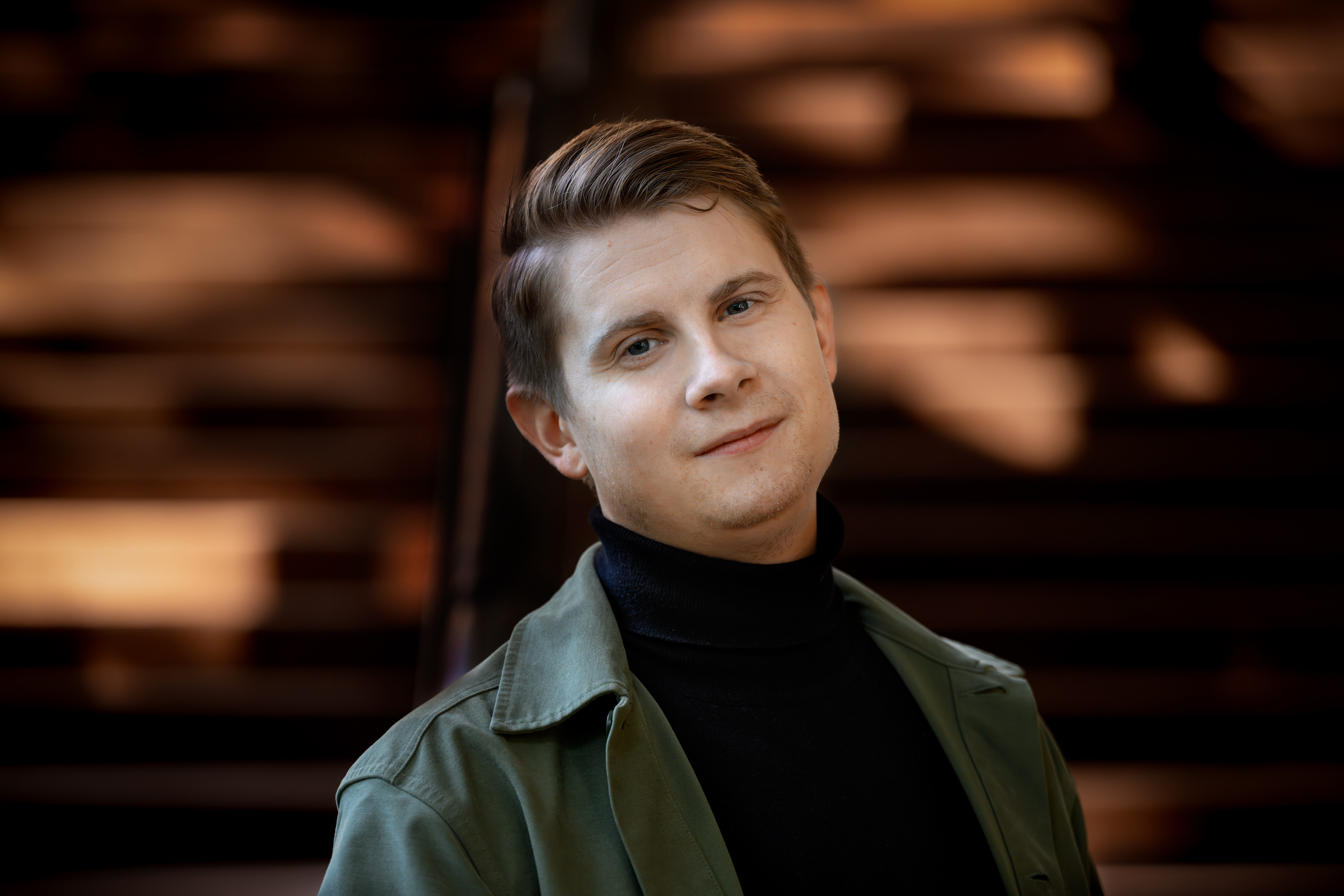 Nuori henkilö, jolla on lyhyet vaaleanruskeat hiukset ja jolla on tumma poolokaulus ja vihreä takki, seisoo puuportaiden ja auringonvalon värittämän taustan edessä. Hän katsoo kameraan hivenen hymyillen.
