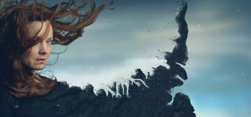 Nainen, jolla on tuulen pyyhkäisemät hiukset, seisoo pilvistä taivasta vasten, ja toinen puoli hänen vartalostaan liukenee karkeaan, tummaan savuiseen tekstuuriin, mikä luo surrealistisen, hajanaisen vaikutelman.