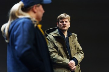 Vihreään takkiin pukeutunut mies seisoo kädet ristissä ja näyttää vakavalta, kun taas siniseen lippikseen ja takkiin pukeutunut henkilö on hämärtynyt etualalla tummaa taustaa vasten.