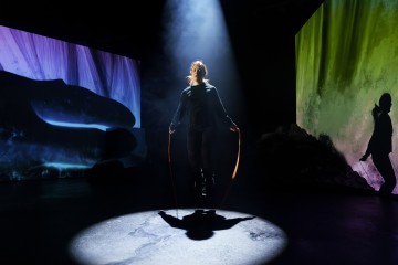 Henkilö hyppää köyttä valokeilan alla pimeällä näyttämöllä, jota ympäröivät värikkäät abstraktit projektiot ja varjot seinillä.