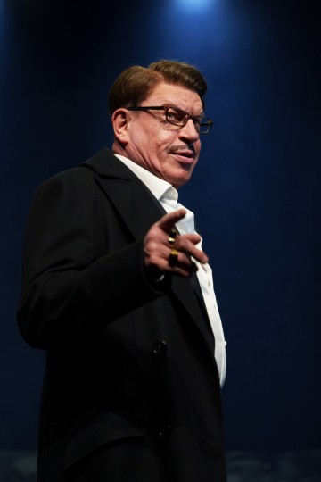 Silmälasipäinen viiksekäs mies, jolla on tumma takki ja valkoinen paita, osoittaa eteenpäin seisoen dramaattisessa näyttämövalaistuksessa tummansinistä taustaa vasten.