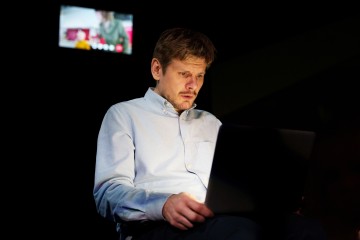 Vaaleansiniseen paitaan pukeutunut mies istuu pimeässä huoneessa ja katsoo keskittyneesti kannettavan tietokoneen näyttöä. Taustalla näkyy epäselvä videopuhelu tai televisioruutu, jossa on toinen henkilö.