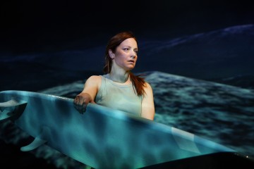 Punatukkainen nainen, jolla on hihaton toppi, seisoo surffilauta kädessään valtamerta vasten dramaattisessa valaistuksessa.