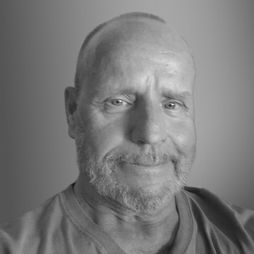 Keski-ikäinen mies, jolla on lyhyet hiukset ja parta, hymyilee lempeästi kameralle. Hänellä on yllään rento paita, ja taustalla on tasainen harmaan sävy. Kuva on mustavalkoinen.