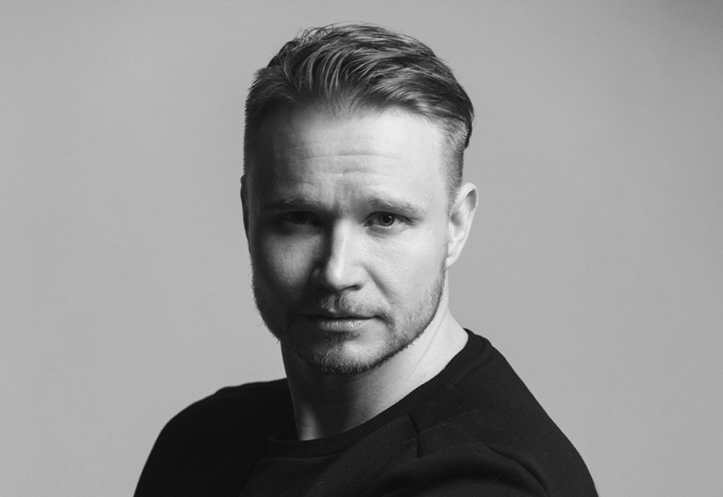 Mies, jolla on lyhyet vaaleat hiukset ja leikattu parta, katsoo vakavana kameraan. Miehellä on tumma paita, ja hänet on kuvattu mustavalkoisessa muotokuvassa tavallista vaaleaa taustaa vasten.