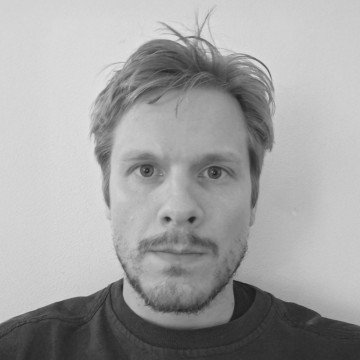 Vaaleat hiukset ja lyhyt parta omaava mies katsoo neutraalilla ilmeellä suoraan kameraan ja seisoo vaaleaa seinää vasten. Kuva on mustavalkoinen.