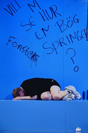 Mustiin vaatteisiin pukeutunut henkilö makaa käpertyneenä sinisellä lattialla sinistä seinää vasten, jossa on ruotsinkielisiä graffiteja, muun muassa sanat Vill ni se hur en bög springer? ja Fönstret. Lähistöllä on vaatteita ja pullo.