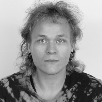 Mustavalkoinen muotokuva henkilöstä, jolla on aaltoilevat, olkapään pituiset hiukset, korvakorut ja solmiopaita, katselee suoraan kameraan neutraalilla ilmeellä.