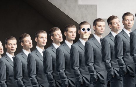Rivi identtisiä harmaapukuisia miehiä seisoo vierekkäin, ja yhdellä keskellä seisovalla miehellä on suuret sarjakuvamaiset silmät ja paperinen suu, mikä erottaa hänet muista.