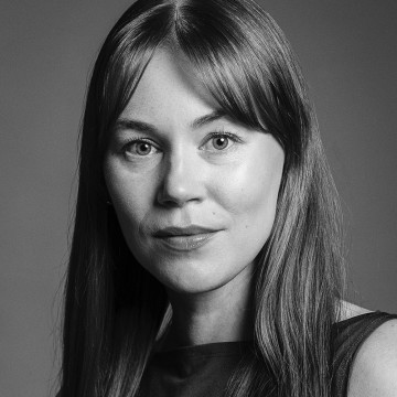 Mustavalkoinen muotokuva nuoresta naisesta, jolla on pitkät suorat hiukset ja otsatukka ja joka katsoo suoraan kameraan neutraali ilme. Hänellä on yllään hihaton toppi ja tausta on tavallinen.