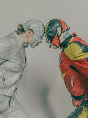 Kaksi futuristiseen asuun pukeutunutta ihmistä, joista toisella on hopeanvärinen kiiltävä puku ja kypärä ja toisella punainen, musta ja keltainen takki ja vastaava kypärä, seisovat kasvotusten ja koskettavat otsaansa tavallista taustaa vasten.